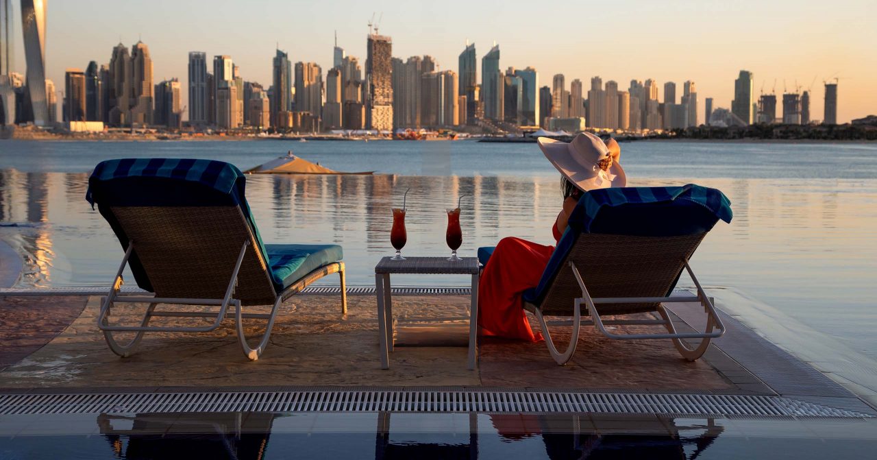 Beautiful beach in Dubai with a lady in red and white hat, 2 sun chairs and an umbrella. In the background is the skyline of Dubai Marina